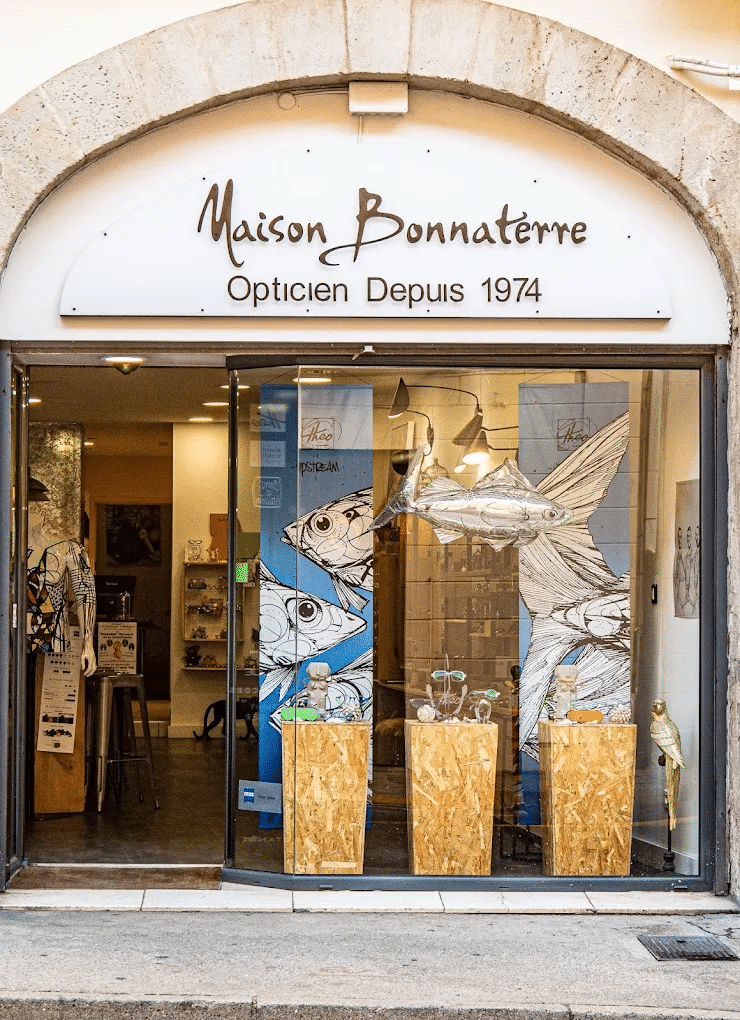 Maison Bonnaterre Opticien : Lunettes & Style Depuis 1974 Devanture de Maison Bonnaterre Opticien Depuis 1974. Vitrine créative avec poisson argenté, illustrations de poissons et lunettes colorées sur socles.
